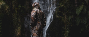 An adult wearing a Selk'bag Realtree® EDGE® Selk'bag sleeping bag suit standing outside near a waterfall